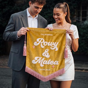 May include: A mustard yellow velvet pennant banner featuring white embroidered text that reads CALIFORNIA, Rowy & Matea, and 25.06.2026. The banner is decorated with two small pink star icons, pink side tassels, and a pink fringe along the bottom edge.