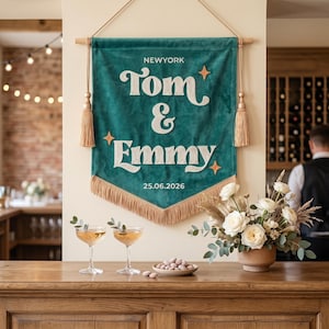 May include: A teal velvet wall banner featuring the white embroidered text NEWYORK, Tom & Emmy, and 25.06.2026. The banner is accented with three orange star icons, two side tassels, and a bottom fringe, all suspended from a wooden rod.