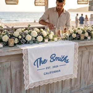 Puede incluir: Un bar de playa con fachada de madera blanca, decorado con conchas marinas. Un camino de mesa blanco con ribete de encaje muestra "The Smiths EST. 2026 CALIFORNIA" en escritura azul. El bar está adornado con rosas blancas y velas.
