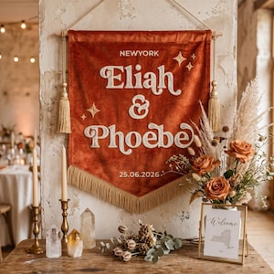 May include: A rust-colored velvet wall banner featuring the embroidered white text NEWYORK, Eliah & Phoebe, and 25.06.2026. The banner is decorated with embroidered stars, gold tassels on the sides, and a gold fringe along the bottom edge.