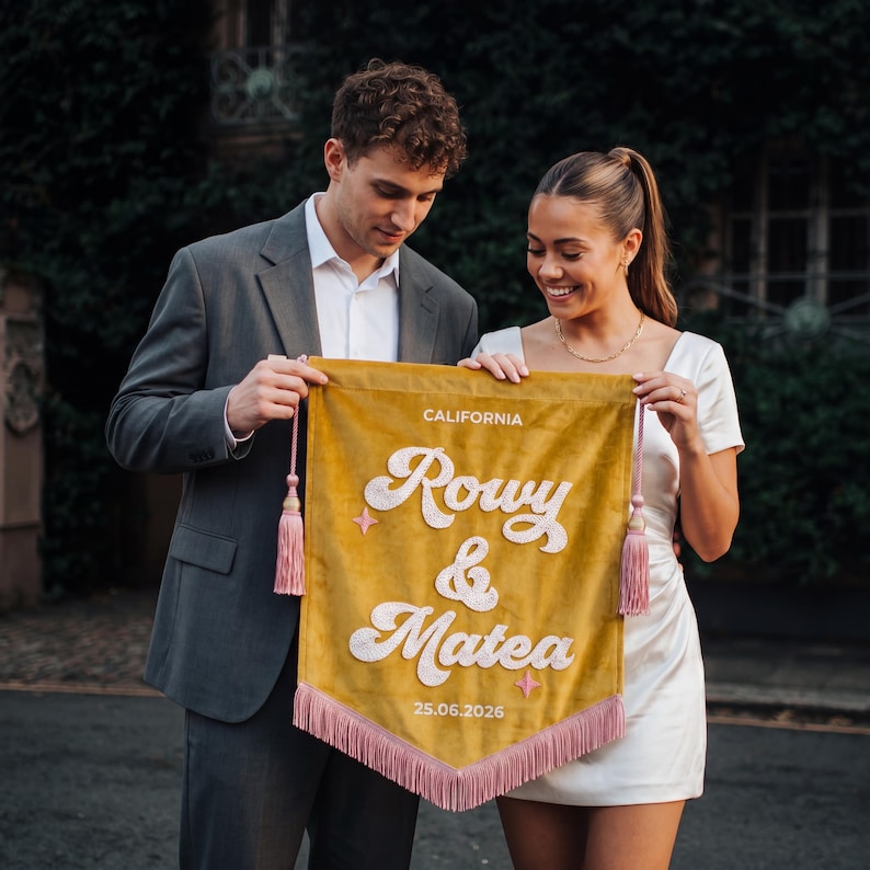May include: A mustard yellow velvet pennant banner featuring white embroidered text that reads CALIFORNIA, Rowy & Matea, and 25.06.2026. The banner is decorated with two small pink star icons, pink side tassels, and a pink fringe along the bottom edge.