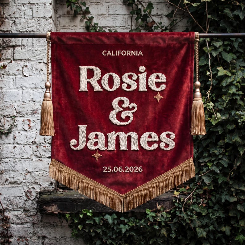 May include: A deep red velvet wall banner featuring the embroidered white text CALIFORNIA, Rosie & James, and 25.06.2026. The banner is accented with two small embroidered stars, gold-colored tassels on each side, and gold fringe at the bottom.