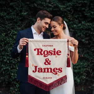 May include: A cream-colored banner with the text "CALIFORNIA Rosie & James 25.06.2026" in red script. The banner has red tassels and fringe. The banner is held by a couple in front of a green hedge.