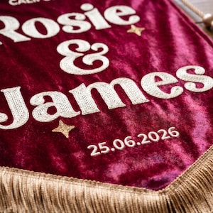 May include: A deep burgundy velvet pennant banner featuring white embroidered text that reads Rosie & James with the date 25.06.2026. The banner is decorated with small gold embroidered stars and finished with gold fringe trim and a hanging cord.