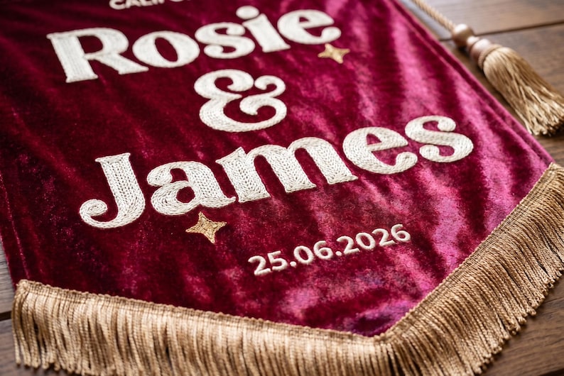 May include: A deep burgundy velvet pennant banner featuring white embroidered text that reads Rosie & James with the date 25.06.2026. The banner is decorated with small gold embroidered stars and finished with gold fringe trim and a hanging cord.