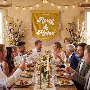 May include: A mustard yellow fabric wall banner featuring the text CALIFORNIA, Rowy & Matea, and 05.26.2026 in white lettering. The banner has a pointed bottom edge with light purple fringe and is suspended by a wooden rod with purple tassels.