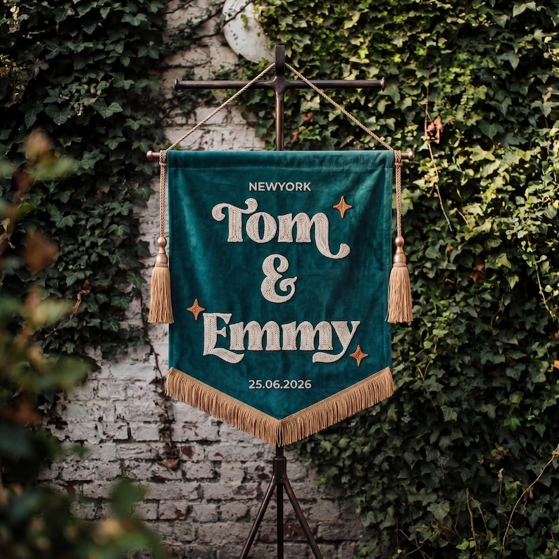 May include: A dark teal velvet pennant banner featuring embroidered cream-colored text that reads NEWYORK, Tom & Emmy, and 25.06.2026. The banner is adorned with four small orange star icons, gold-colored tassels on each side, and gold fringe at the bottom.