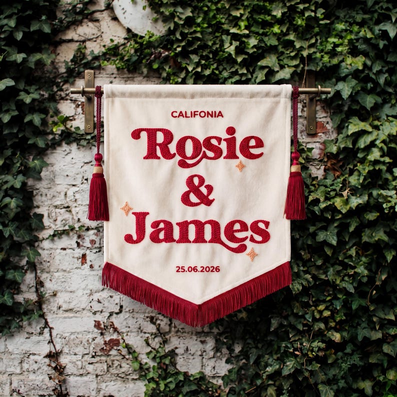 May include: A cream-colored fabric banner featuring the embroidered red text "CALIFONIA", "Rosie & James", and the date "25.06.2026". The banner includes red tassels on both sides, a red fringe at the bottom, and small embroidered star accents.