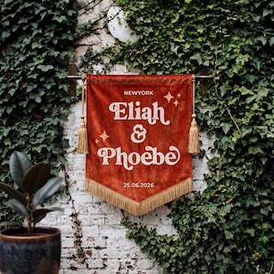 May include: A rust-colored velvet wall banner featuring the text NEWYORK, Eliah & Phoebe, and 25.06.2026 in white lettering. The banner is accented with white star icons, gold-toned tassels on each side, and a gold-toned fringe along the bottom edge.