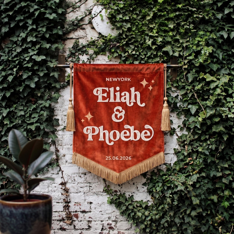 May include: A rust-colored velvet wall banner featuring the text NEWYORK, Eliah & Phoebe, and 25.06.2026 in white lettering. The banner is accented with white star icons, gold-toned tassels on each side, and a gold-toned fringe along the bottom edge.