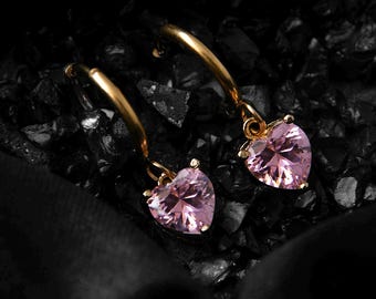 May include: A pair of gold hoop earrings with pink heart-shaped charms. The earrings are dangling and the hearts are accented with sparkling rhinestones.