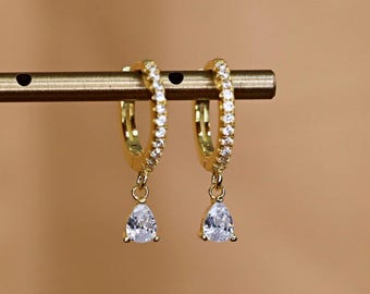 May include: A pair of gold hoop earrings with a teardrop-shaped, clear gemstone pendant. The hoops are encrusted with small, clear gemstones. The earrings are displayed on a gold-colored bar against a warm, neutral background.