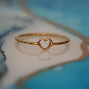 Op de afbeelding: Een delicate gouden ring met een hartvormige uitsparing. De ring heeft een dunne band en een gepolijste afwerking, die het licht reflecteert. Het hart is gecentreerd op de band, waardoor een eenvoudig maar elegant ontwerp ontstaat. De ring is een sieraad.