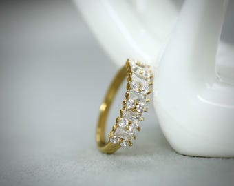 May include: A gold ring with a band of alternating baguette and round-cut clear gemstones. The ring is displayed against a white background, showcasing its sparkle and design. The ring is a piece of jewelry.