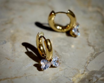 May include: Gold-colored hoop earrings with a bow-shaped, clear gemstone charm. The earrings are made of a shiny, reflective metal and are displayed on a marble surface. The earrings are a classic jewelry item.