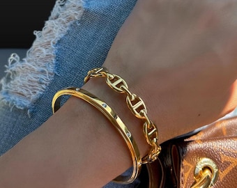 May include: Two gold bracelets on a wrist. One bracelet is a thin bangle with small clear stones. The other bracelet is a thicker chain with a mariner link design.