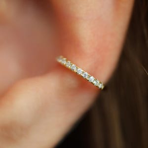 May include: A close-up of a gold-colored ring with a row of small, clear gemstones. The ring is worn on an ear, with the skin in the background slightly blurred. The ring's design is simple and elegant, with the gemstones adding sparkle.