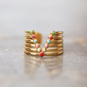 May include: A gold-colored ring with a multi-layered design. The ring features a series of parallel bands, with a V-shaped row of colorful, round accents in green, pink, blue, white, and red. The ring is set against a neutral background.