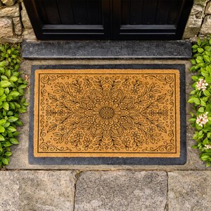 K&ouml;nnte beinhalten: Eine rechteckige Fu&szlig;matte mit einem floralen Mandala-Design. Die Matte besteht aus einem nat&uuml;rlichen, beigefarbenen Material mit einem schwarzen Rand. Das florale Muster ist schwarz und zeigt eine zentrale Blume mit umliegenden Bl&auml;ttern und kleineren Bl&uuml;ten. Die Fu&szlig;matte liegt vor einer schwarzen T&uuml;r.