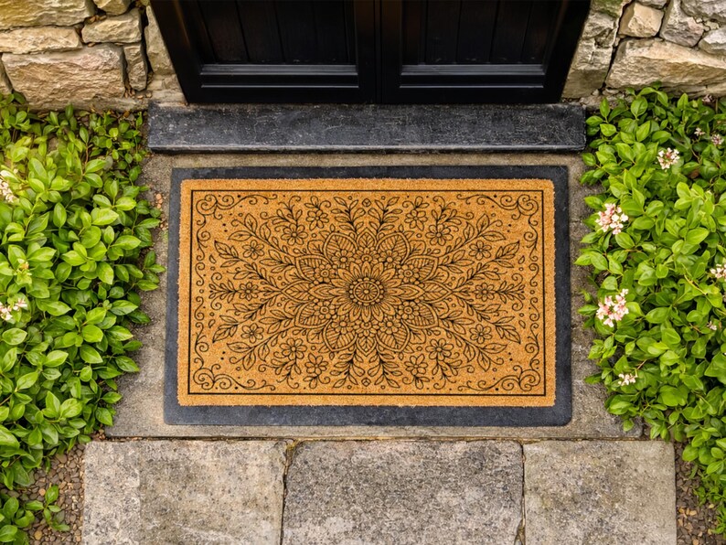 K&ouml;nnte beinhalten: Eine rechteckige Fu&szlig;matte mit einem floralen Mandala-Design. Die Matte besteht aus einem nat&uuml;rlichen, beigefarbenen Material mit einem schwarzen Rand. Das florale Muster ist schwarz und zeigt eine zentrale Blume mit umliegenden Bl&auml;ttern und kleineren Bl&uuml;ten. Die Fu&szlig;matte liegt vor einer schwarzen T&uuml;r.