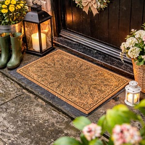 K&ouml;nnte beinhalten: Eine rechteckige Fu&szlig;matte mit detailliertem floralem Design vor einer T&uuml;r. Die Matte ist braun mit schwarzen Details. Eine schwarze Laterne mit einer brennenden Kerze, gr&uuml;ne Stiefel und ein Blumenkorb befinden sich in der N&auml;he.