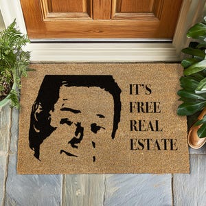 May include: A rectangular doormat made of natural fibers. The mat features a black silhouette of a person's face and the phrase "IT'S FREE REAL ESTATE" in black, bold letters. The mat is placed in front of a wooden door.