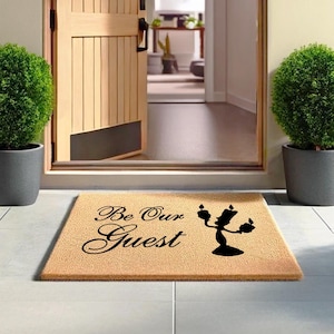May include: A brown doormat with the text "Be Our Guest" in black cursive font. A black silhouette of Lumiere, the candelabra from Beauty and the Beast, is on the right side of the mat.