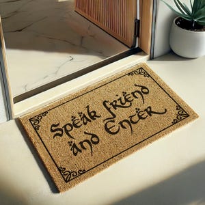 May include: A rectangular doormat with the words "Speak Friend and Enter" in a decorative font. The mat is a natural tan colour with black lettering and border. Ornate flourishes are in each corner. The mat is placed in a doorway.
