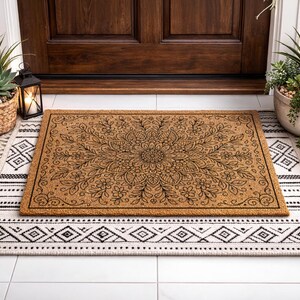 K&ouml;nnte beinhalten: Eine rechteckige Kokos-Fu&szlig;matte mit einem schwarzen Blumen-Mandala-Design liegt auf einem schwarz-wei&szlig; gemusterten Teppich. Die Fu&szlig;matte ist braun und der Teppich hat ein geometrisches Muster. Die Szene spielt sich vor einer Holzt&uuml;r ab.