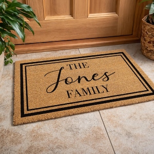 Könnte beinhalten: Eine rechteckige Fußmatte mit dem Text "THE Jones FAMILY" in schwarzer Schrift. Die Matte besteht aus einem hellbraunen Material mit schwarzem Rand. Die Matte liegt vor einer Holztür.