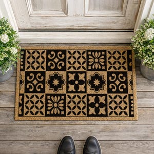 May include: Rectangular doormat with a geometric black and tan pattern. The design features alternating squares with floral and scroll motifs. The mat is made of a natural fiber material and is placed in front of a white door.
