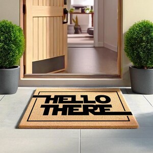 Pode incluir: Um tapete de coco castanho com uma borda preta e o texto "HELLO THERE" em letras pretas.