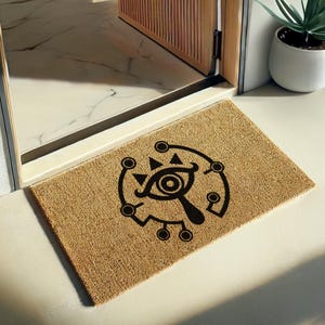 Op de afbeelding: Een rechthoekige deurmat van natuurlijke vezels, met een zwart grafisch ontwerp van een oog in een cirkelvormig en geometrisch patroon. De mat ligt voor een open houten deur.