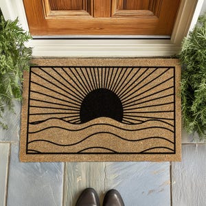 Könnte beinhalten: Eine rechteckige Fußmatte mit einem schwarzen Sonnen- und Wellenmuster auf beigem Hintergrund. Die Sonne befindet sich in der Mitte, mit Strahlen, die sich nach außen erstrecken. Die Wellen befinden sich unter der Sonne. Die Matte liegt vor einer Holztür.