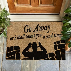 Op de afbeelding: Een bruine kokosvezel deurmat met de tekst "Go Away or I shall taunt you a 2nd time" in zwarte letters. Een zwart silhouet van een persoon met zijn handen over zijn oren bevindt zich in het midden van de mat.