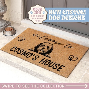 Op de afbeelding: Een rechthoekige deurmat met de tekst "welcome to COSMO'S HOUSE" en een hondenillustratie. De mat is bruin met zwarte tekst en een hondenafbeelding. De tekst "OVER 100 DOG BREEDS TO CHOOSE FROM" is ook zichtbaar.