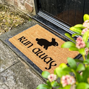 May include: A rectangular coir doormat with the words "run away" and a black rabbit silhouette. The text is in a playful font. The mat is placed in front of a black door, on a stone step.