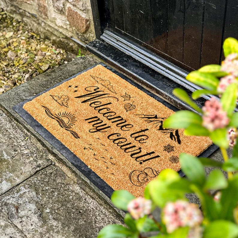 May include: A rectangular doormat made of natural fibers, featuring the phrase "Welcome to my court!" in elegant black script. The design includes illustrations of a sun, clouds, leaves, and other decorative elements. The mat is placed in front of a black door.