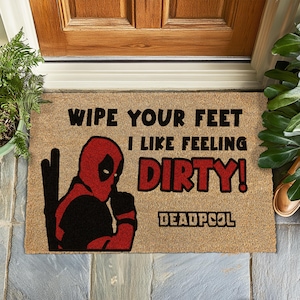Könnte beinhalten: Eine braune Kokosfaser-Fußmatte mit dem Text "Wipe your feet I like feeling dirty! Deadpool" in schwarzer Schrift. Links auf der Matte befindet sich eine rote und schwarze Illustration von Deadpool.