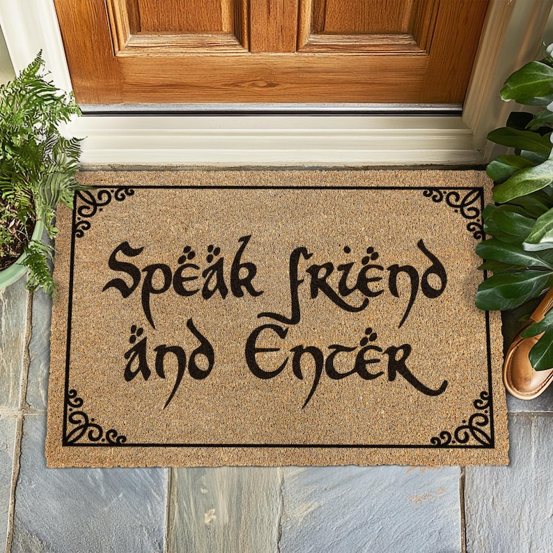 May include: A rectangular coir doormat with the text "Speak friend and Enter" in black, stylized lettering. The mat has a black border with decorative corner designs. The mat is placed in front of a wooden door.