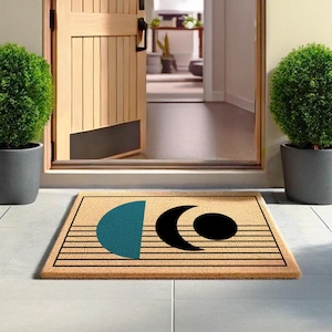 Può includere: Un tappetino per porta in fibra di cocco marrone con un design geometrico che presenta un semicerchio blu turchese, una mezzaluna nera e un cerchio nero. Il tappetino ha linee nere che corrono orizzontalmente attraverso di esso.
