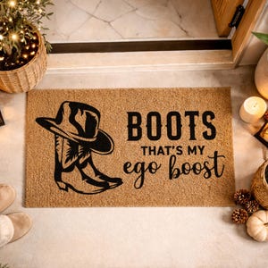 Puede incluir: Una alfombra rectangular con el texto "BOOTS THAT'S MY ego boost" y un gráfico de un sombrero y botas de vaquero. La alfombra es marrón con texto y diseño en negro. Elementos decorativos incluyen velas, plantas y una calabaza pequeña.