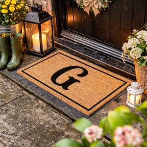 May include: A rectangular doormat with the letter "G" in black, framed by a black border, sits on a stone surface. The mat is in front of a black door, with a lantern and potted plants nearby. Green rain boots are also visible.