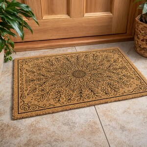 K&ouml;nnte beinhalten: Eine rechteckige Fu&szlig;matte mit einem floralen Mandala-Design. Die Matte besteht aus einem nat&uuml;rlichen, beigefarbenen Material mit einem schwarzen Rand und einem schwarzen Blumenmuster. Die Matte liegt vor einer Holzt&uuml;r.
