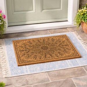 K&ouml;nnte beinhalten: Eine rechteckige Fu&szlig;matte mit einem floralen Mandala-Design in Braun- und Schwarzt&ouml;nen. Die Fu&szlig;matte liegt auf einem hellblauen Teppich mit wei&szlig;em Fransen. Die Szene spielt sich vor einer gr&uuml;nen T&uuml;r ab, mit Topfpflanzen auf beiden Seiten.