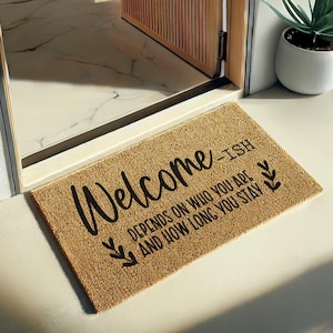 Pode incluir: Um capacho retangular feito de fibras naturais, com a palavra "Welcome" em fonte cursiva. O texto "-ish DEPENDS ON WHO YOU ARE AND HOW LONG YOU STAY" também está impresso no tapete, juntamente com detalhes decorativos de folhas.