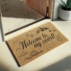 May include: A rectangular doormat made of natural fibers, featuring the phrase "Welcome to my court!" in elegant black script. The design includes mountain, sun, cloud, and leaf illustrations. The mat is placed in front of a doorway.