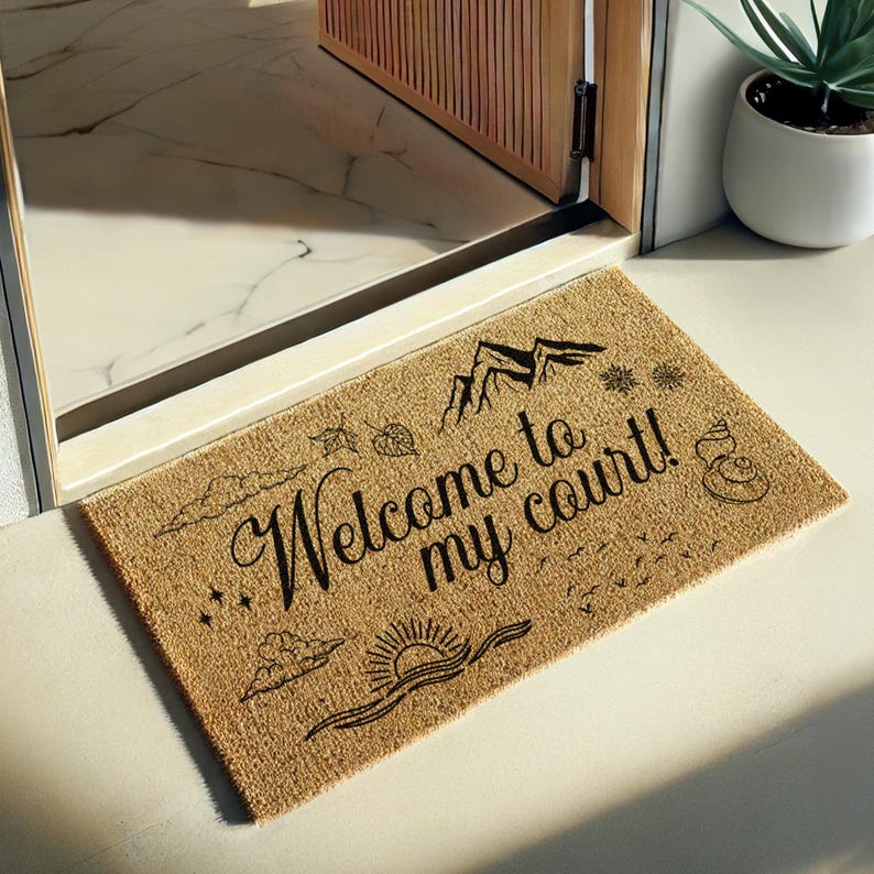 May include: A rectangular doormat made of natural fibers, featuring the phrase "Welcome to my court!" in elegant black script. The design includes mountain, sun, cloud, and leaf illustrations. The mat is placed in front of a doorway.