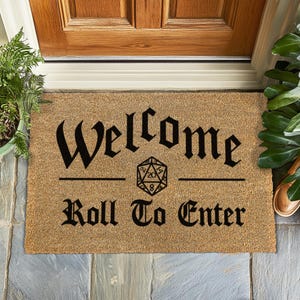 Puede incluir: Una alfombra de coco marrón con el texto "Welcome Roll To Enter" en negro. Un dado negro y blanco de 20 caras está centrado entre las dos líneas de texto.