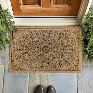 A top-down view of a rectangular tan coir doormat featuring an elaborate black botanical mandala design. The pattern radiates from a central flower, surrounded by layers of detailed petals, leaves, and smaller blossoms that fill the entire mat. Decorative scrollwork flourishes accent the four corners, all enclosed by a thin black rectangular border.